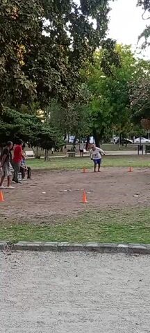 The video shows a group of people in a park setting. There are several mature trees and patches of grass, some of which is covered in gravel. In the center, there's a dirt area marked with orange cones and what looks like discs or tiles scattered on the ground. To the left of the dirt area, there are four men: three standing and one seated on a bench. In the background, there are other people sitting on benches and walking around, and some cars parked on the street in the distance. One person walks into the dirt area from the right and stands near the cones and discs.