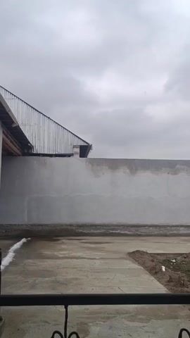 The video shows a view from a balcony or porch, looking out at a walled-in yard under a cloudy sky. The wall is made of concrete and has dark patches or stains on it. There is a building to the left with a corrugated metal roof. The ground is mostly concrete, with some patches of dirt and some small piles of snow. A dark, wrought-iron railing is visible at the bottom of the frame. The camera starts out focused on the scene and then goes slightly out of focus.