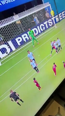 The video shows a soccer match on a large screen. A goalkeeper in a green uniform is diving to his left in front of a net. Several players in light blue and red uniforms are on the field. The net has a large "PREDICTS" advertisement on it, with "WORLD CUP 2026" visible above it. Spectators are seated in the background. The camera is angled from the side and slightly below the screen.
