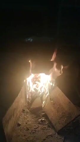 A small, brightly burning fire is centered in the frame. The fire is contained within a V-shaped structure made of two pieces of wood resting on a dirt or ash-covered surface. Flames lick upwards, casting a warm glow on the surroundings. The background is dark and indistinct, with faint hints of objects and light sources suggesting an outdoor or dimly lit indoor setting. The camera appears to be held at a slight downward angle, focusing on the fire.