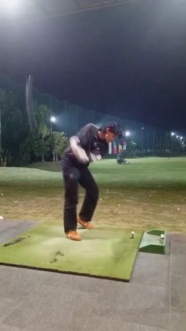 A person is practicing golf at a driving range at night. The person is wearing a black long-sleeved shirt, dark jeans, and orange shoes. The person is standing on a green mat and is swinging a golf club. There are golf balls on the ground and in a basket. There are lights and flags in the background.