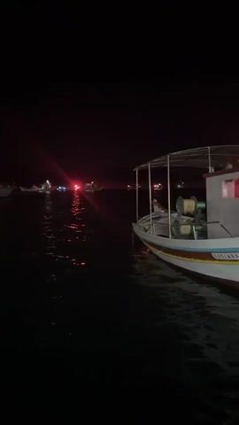 The video shows a body of water at night. A small boat is visible in the foreground on the right, with a canopy overhead and equipment on its deck. In the distance, several other boats are present, with one in the center emitting a bright red flashing light and other lights in green and white. The water reflects the lights, creating streaks of color on the dark surface. The overall environment appears to be a harbor or a body of water with other vessels.