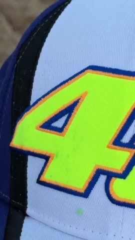 The video displays a close-up, angled view of a light blue baseball cap. Prominently stitched onto the cap are large, bold letters in a neon yellow-green color. These letters are outlined with a dark blue thread and then further bordered by a thin orange thread. The texture of the cap's fabric is visible, showing the weave of the material. The lighting appears to be even, highlighting the vibrant colors of the stitching.