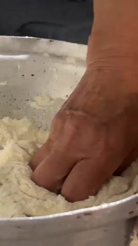 A close-up shot of a light-skinned hand kneading a dough in a silver bowl. The hand is covered in the dough, which is a light cream color. The bowl is also speckled with the dough. The background is out of focus, appearing as a dark, blurred area.