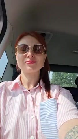 A woman with red hair and sunglasses is in the driver's seat of a car, wearing a pink and white striped shirt. A younger person with curly hair is in the passenger seat behind her, smiling and making peace signs. The interior of the car is visible, with bags and luggage in the back. The surroundings outside the car appear to be a residential area with a building and trees.