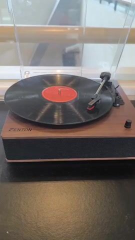 A video shows a Fenton record player playing a vinyl record with a red center label. The record player is a wood-look finish with a black fabric-covered base and a clear plastic dust cover that is in the open position. The tonearm is in place over the spinning record. The record player is on a black surface, possibly a table, in front of a blurred-out background that looks like a window or glass barrier with wooden railings.  The audio contains music.