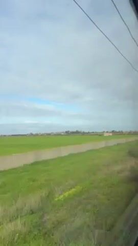 The video shows a view from a moving train. The train is traveling through a rural area with green fields. In the distance, there is a small town or village. There are utility poles and electrical wires visible outside the window. The sky is cloudy with patches of blue. The video appears to be taken during the daytime.