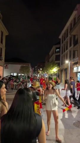 The video shows a street scene at night. A group of people, many wearing red and yellow, possibly in celebration of a sporting event, are walking down a street lined with buildings. The people are diverse in age and gender. Some are carrying Spanish flags and noise-making items. The street is paved and illuminated by streetlights and lights from shops and restaurants. There are tables and chairs set up outside some of the establishments. The overall atmosphere appears festive and energetic.
