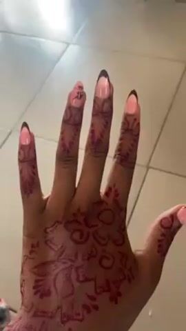 A dark-skinned hand with henna tattoos on the fingers and back of the hand is the focus. The fingernails are polished with a pink color and have black tips. The henna designs are intricate, with flower patterns, swirls, and linear elements. The hand is shown on a background of square, light-colored floor tiles. The hand rotates to display the tattoos from different angles. Music plays in the background.