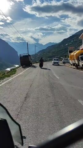 The video shows a motorcycle riding on a paved road in a mountainous region. The road curves to the right and ascends a hill. On the left side of the road is a steep drop to a valley with a river and a small settlement. In the background, there are tall mountains with snow-capped peaks. The sky is partly cloudy with the sun shining through. There are other vehicles on the road, including a truck parked on the left and a car driving in the same direction as the motorcycle. The motorcycle is ridden by a person wearing a helmet and dark clothing. The video is shot from the perspective of another motorcycle or vehicle, with a portion of the windshield visible in the foreground. The overall impression is of a scenic road trip through a beautiful natural landscape.