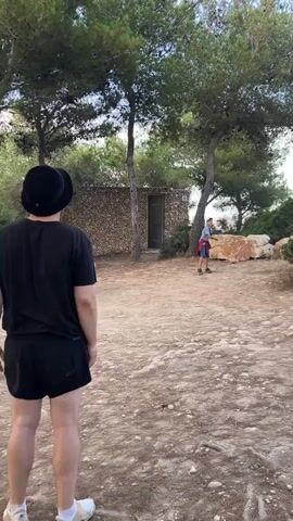 A person wearing a black t-shirt, black shorts, a black bucket hat, and white sneakers is standing in a dirt area with trees and a small stone building in the background. Another person, wearing a red shirt, blue shorts, and an orange hat, is in the distance. The first person is facing away from the camera, then turns to look at the camera and smiles. There is a small orange object on the ground in front of the first person.