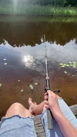 A first-person perspective video shows a person fishing from a wooden dock. The person's legs, clad in denim shorts, and bare feet are visible in the foreground. They are holding a fishing rod with a reel, casting into a dark body of water with lily pads. The sun is shining brightly, reflecting off the water's surface, and a line of green reeds lines the opposite bank of the water. The video is shot during daylight hours, indicating an outdoor setting.