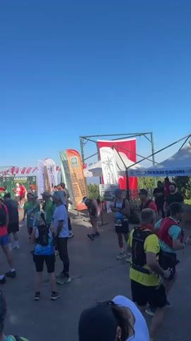 The video shows a gathering of people outdoors on a sunny day, likely for a race or event. In the foreground, a group of people are milling around, some wearing athletic clothing and backpacks. Behind them, a stage is set up with a banner that reads "OSMAN UL TRA TRAIL HOŞ GELDİNİZ," suggesting it's a trail running event. Several flags and banners are visible, including what appears to be a Turkish flag. People are standing, talking, and some are stretching or preparing. A tall cell tower is visible in the background, as are trees and buildings. The overall atmosphere appears to be one of anticipation and preparation for an outdoor activity.