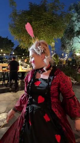 An outdoor evening scene shows a woman dressed as the Queen of Hearts, striking a pose for the camera. She wears a black dress with red heart cutouts and a long red and black patterned robe. Her costume includes a black choker, a gold crown with a large red heart on top, and heart-shaped makeup on her cheeks and lips. She holds a gold and red scepter in her right hand.

In the background, other people are visible, mostly out of focus, suggesting an event or gathering. The setting is illuminated by numerous string lights strung between trees, indicating it's nighttime or dusk. The ground appears to be a paved or tiled area in a garden or event space. The overall atmosphere suggests a festive or themed event. No text is visible in the image. The location is Nir Eliyahu, Israel.