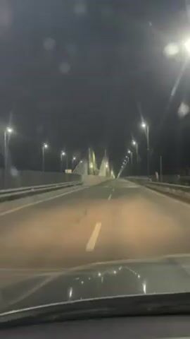 The video shows a nighttime drive across a bridge. The view is from inside the car, looking forward. The bridge is lit by street lamps that line the sides. In the distance, the bridge's towers are visible, illuminated with white, yellow, and purple lights. The road is marked with a solid white line. There are water spots on the windshield, and the dashboard is visible at the bottom of the frame.