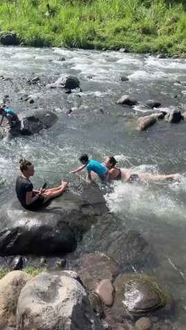 Two women and two children are playing in a shallow, rocky river. One woman sits on a rock on the left, holding a phone. The other woman is in the water with two children. One child, wearing a blue shirt, is being held by the woman, who is lying on her back. Another child, wearing a rash guard and shorts, stands in the water on the right, looking down. The river is flowing with visible ripples and rocks are scattered throughout. Lush green vegetation lines the bank in the background.