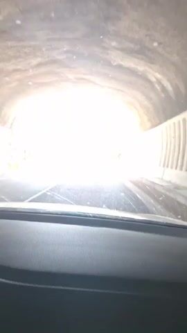 The video begins with a view from inside a vehicle exiting a tunnel. The tunnel exit is brightly lit, with the road ahead appearing washed out by the sunlight. As the vehicle emerges, the camera captures a winding road with a mountain landscape. There are warning signs indicating sharp curves. The scenery transitions to a view of a mountainous terrain with lush greenery and scattered rocks under a bright, cloudy sky.