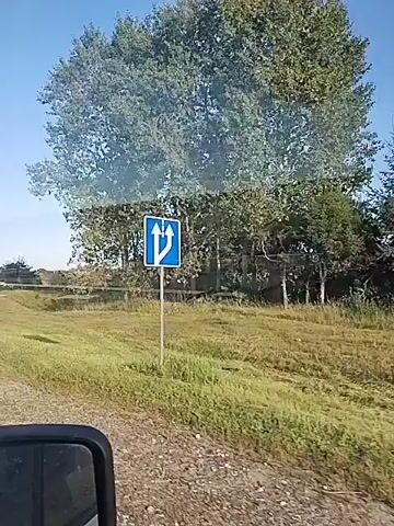 The video is a first-person view from a vehicle traveling on a road. The road is paved with a white line marking the edge. To the right of the road is a grassy shoulder and then a field of cut grass. Beyond the field, there are trees and some buildings that appear to be houses. The sky is clear and blue. In the distance, there are utility poles and signs. A side mirror is visible in the lower left corner of the frame. The video captures a daytime scene with good lighting.