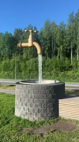 A water feature sculpture resembling a large, golden faucet is shown in this video. The faucet is positioned above a circular stone well, with water continuously flowing from the spout into the well. The well is constructed of gray, rectangular stone blocks and has a concrete rim. Surrounding the well is green grass. In the background, a dense forest with tall trees forms a backdrop under a clear blue sky. To the right of the well, a wooden bench-like structure is partially visible. The video appears to be filmed outdoors during daylight.