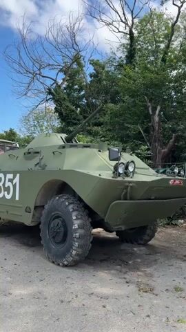The video shows a military armored personnel carrier with a cannon mounted on its turret. The vehicle is painted olive green and has the number "251" on its side. It appears to be parked on a dirt or gravel surface with trees and foliage in the background. The sky is blue with some clouds. The overall impression is that of a military vehicle in an outdoor setting.