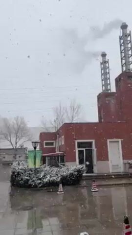 The video shows a snowy day. The camera pans across a red brick building with two smokestacks emitting smoke. Snow falls heavily, accumulating on parked cars and surrounding bushes. The sky is overcast and the visibility is reduced due to the snowfall. In the background, apartment buildings can be seen. The overall environment suggests an urban or industrial area during winter.