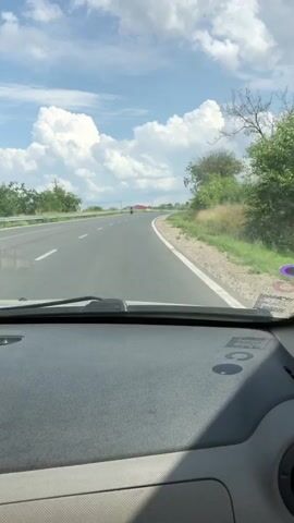 This video shows a first-person perspective from inside a vehicle, likely a car, as it travels along a paved road. The road appears to be a highway or a major route, with clear lane markings and a white line on the right shoulder. The scene is set during the daytime, with a bright, partly cloudy sky. The weather appears to be warm, with lush green vegetation on the sides of the road, including trees and tall grasses.

In the distance, a lone motorcyclist can be seen riding ahead on the road. Beyond the road, the landscape is relatively flat with some scattered buildings and more trees. The overall mood is peaceful and scenic, suggesting a pleasant journey through a rural or semi-rural area.

Visible on the dashboard of the car are some smudges and possibly some stickers or markings. A purple circular sticker is visible on the lower right side of the windshield. The location is identified as Bulboaca, Moldova. No specific emotional context is conveyed beyond the general feeling of a journey.