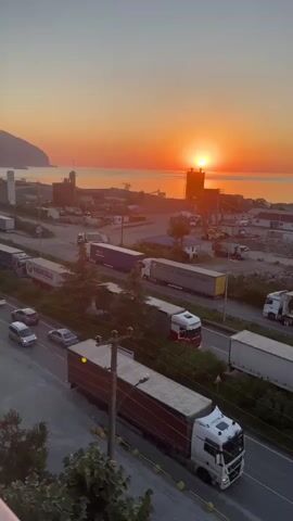 The video captures a sunrise over a body of water, likely the sea, with a coastal landscape visible in the distance. In the foreground, there is a busy road with multiple lanes of traffic, including several large semi-trucks and some smaller cars. The trucks appear to be parked or moving slowly, suggesting a possible traffic jam or a waiting area. The environment seems to be industrial or a port area, with some buildings and structures visible near the water's edge. The sky is painted with warm orange and yellow hues from the rising sun, creating a serene yet active atmosphere. The camera is positioned at an elevated angle, offering a panoramic view of the scene.