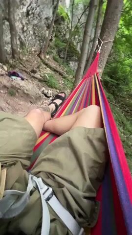 The video begins with a first-person view from a hammock, showing the legs and torso of a person. The hammock is brightly colored with red, yellow, and green stripes. Behind the hammock, a sheer rock face is visible, with trees and foliage in the background. The camera then pans slightly to the left, revealing two people rock climbing on the rock face. One climber, wearing a green shirt and shorts, is higher up on the wall, while another climber, a female wearing a white shirt, helmet, and climbing harness, is standing at the base of the rock face. Ropes and climbing gear are scattered on the ground. The environment appears to be a wooded area with a rocky terrain.