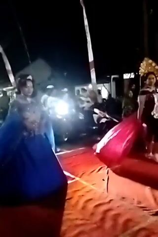 At night, a woman in a blue dress with intricate beading is standing on a red carpet. She is looking to her left. To her right, a person in a grey shirt and dark pants is walking away from the camera. Behind them, several other people are visible, some holding what appear to be flags or poles. There are also motorcycles parked in the background. The scene seems to be outdoors, possibly a street or an event venue. The lighting is dim, with artificial lights illuminating the area.