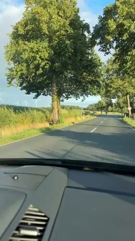 The video shows a first-person perspective of a car driving down a paved road lined with trees on both sides. The road has a white dashed line down the center. On the left side of the road, there is a field with tall grass and several wind turbines in the distance. The sky is blue with scattered white clouds. The interior of the car, including the dashboard, is visible in the foreground. The overall scene depicts a rural landscape with a clear sky and good visibility.