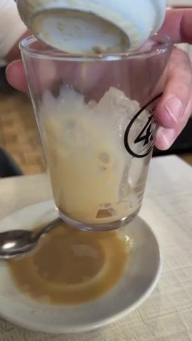 A close-up shot shows a clear glass with ice and a beige liquid being poured into it. The glass has a logo with the number 40. A small white pitcher is being held above the glass, from which the liquid is being poured. Below the glass, a white saucer holds a small amount of the same beige liquid, and a silver spoon rests on the saucer. The background is blurred but appears to be a table setting.