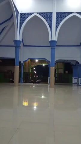 The video shows the interior of a building with a high ceiling supported by blue and tan pillars. The walls and arches are painted white with blue decorative trim. The floor is a highly reflective white tile. In the background, an illuminated archway leads to an outdoor area where motorcycles and people are visible. The lighting suggests it is evening or night.