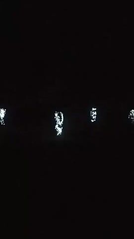 A dark scene shows several blurred clusters of white lights in the distance, appearing to be reflections on water. The lights are spread out horizontally across the frame and are the only discernible objects in the shot.