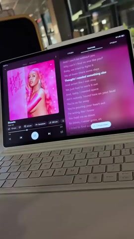 The video shows a close-up of a tablet computer with a white keyboard attached, positioned on a table. The tablet screen displays a music player interface, showing album art with a woman in pink, the song title "Streets" by Doja Cat, and scrolling lyrics in English. The current lyric highlighted on the screen is "And acted like I was okay". The music player controls and song progress bar are visible at the bottom of the screen. The background is blurred, suggesting an indoor environment like a cafe or restaurant. The lighting is soft, and the overall scene appears casual and relaxed.
