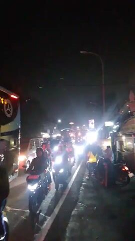 A nighttime scene captures a congested road filled with numerous motorcycles and a large bus. The motorcycles, many with their headlights illuminated, are lined up in multiple lanes, indicating slow-moving or stopped traffic. Some riders are wearing helmets, and several motorcycles appear to carry passengers. The bus, on the left side of the frame, has its brake lights illuminated. Streetlights and building lights contribute to the overall illumination of the scene, creating bright spots and reflections on the wet road surface. Several individuals are visible standing on the side of the road, observing the traffic. The environment appears to be an urban or semi-urban area with buildings lining one side of the road. The overall impression is one of heavy traffic congestion at night.