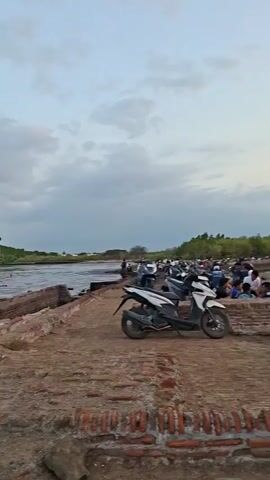 A person stands on a brick path overlooking a body of water. In the background, there are trees and a cloudy sky. To the right, several motorcycles are parked. The person is wearing a grey t-shirt and beige shorts. The motorcycles are various models and colors, some appear to be covered with tarps or bags. The environment appears to be a coastal or riverside area with some visible debris.