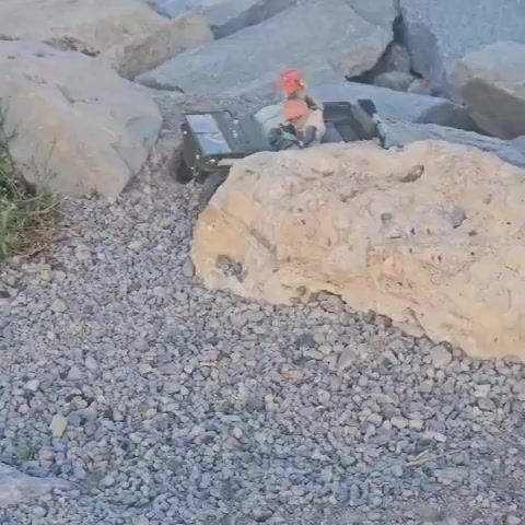 An army-green remote control (RC) jeep, carrying two small action figures, navigates a rocky, gravelly outdoor terrain. One figure has a red head covering, while the other has a beige or white head covering. The ground is covered in small grey and white pebbles, interspersed with larger, light-colored boulders and sparse green vegetation. The jeep drives across the uneven ground, moving from the upper-left of the frame towards the lower-right before turning and proceeding to the left. On the rear of the jeep, white text resembling a serial number, "2011152879," is visible. The scene is well-lit by natural daylight, indicating clear weather. The setting suggests a natural or semi-natural outdoor area, consistent with a rocky coastal or inland environment found in Savona, Italy.