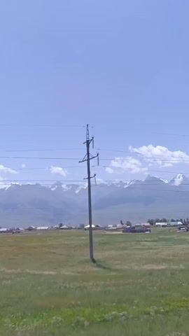 The video shows a wide shot of a rural landscape under a clear blue sky. In the foreground, there is a field of green grass. In the middle ground, there are several scattered buildings, some appearing to be houses and others possibly under construction. Beyond the buildings, a range of majestic mountains looms in the distance, some with snow-capped peaks. Power lines stretch across the frame in the upper portion of the video. The perspective suggests the camera is moving, as evidenced by the slight shift in the visible landscape between frames. There are no people or animals visible in the footage.