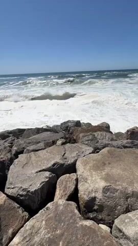 This media file is a vertical video capturing a dynamic ocean scene at Torreira, Portugal. The primary subjects are the crashing waves of the Atlantic Ocean and the rocky shoreline in the foreground. Large, weathered boulders dominate the lower portion of the frame, forming a natural barrier against the sea.

The activity taking place is the relentless motion of the ocean. Waves are seen building, cresting, and breaking against the rocks, creating a white froth and spray. The water appears energetic, indicative of a moderately rough sea. The sky above is a clear, vibrant blue, suggesting a bright, sunny day. The lighting suggests it is daytime, likely mid-morning to early afternoon, given the strong sunlight casting distinct shadows on the rocks and illuminating the water.

The scene is a coastal landscape. The vast expanse of the ocean stretches to the horizon, meeting the clear sky. The location cues from "Torreira, Portugal" point to a specific coastal town in Portugal, known for its beaches and proximity to the Atlantic. There are no people visible in the video, emphasizing the raw power and beauty of nature. The overall emotion conveyed is one of natural grandeur and the powerful force of the sea. No visible text is present in the media file.