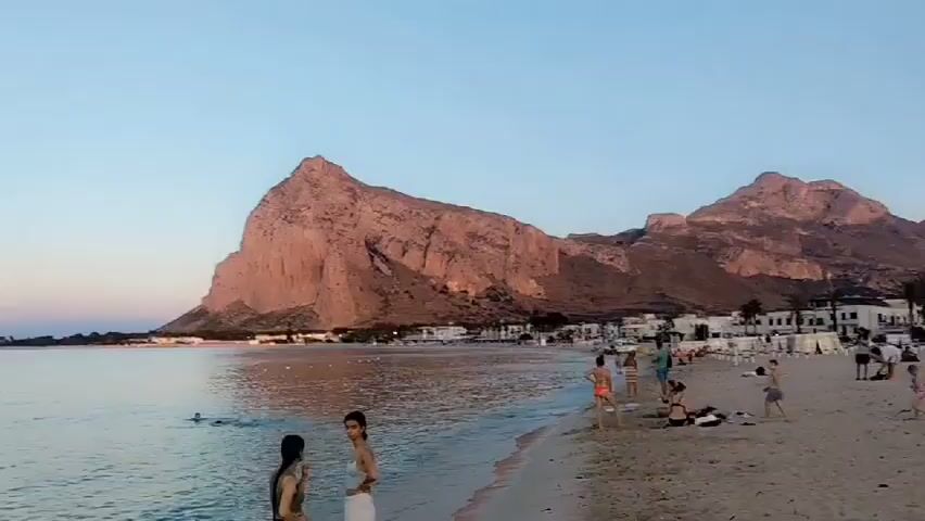 This media file captures a tranquil scene at San Vito Lo Capo, Italy, likely during a summer evening. The setting is a picturesque beach with a calm sea and a dramatic, rocky mountain dominating the background. The time of day is evident from the soft, warm hues of the sunset casting a gentle glow over the sky and reflecting on the water.

Several people are present, enjoying the beach and the sea. Two women are standing near the water's edge in the foreground, appearing relaxed. Further out in the water, a few individuals are swimming or wading, indicating a pleasant temperature and a leisurely atmosphere. On the sandy shore to the right, more beachgoers are visible, some sunbathing or relaxing.

Numerous sailboats and catamarans are anchored in the bay, adding to the serene maritime ambiance. Their masts stand tall against the fading light, suggesting that the day's sailing activities are winding down. A distant coastline is visible on the left, with a structure that might be a pier or a breakwater.

The overall mood is peaceful and idyllic. The clear, soft sky and the gentle lapping of waves create a sense of calm and relaxation. The location cues, particularly the distinctive mountain, firmly place the scene in San Vito Lo Capo, a popular coastal town in Sicily known for its beautiful beaches. There is no visible text in the media file.