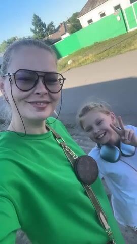 A selfie video is taken outdoors on a sunny day. Two females are visible in the foreground. The female on the left is wearing large black-rimmed glasses and a bright green sweatshirt. She is smiling at the camera. A thin black cord from her glasses dangles down her chest, and a small brown round bag with a monogram pattern is attached to a strap across her shoulder. The female on the right is wearing a white t-shirt and large blue headphones around her neck. She is smiling and looking at the camera, then makes a pouting face. In the background, there is a paved road, a grassy area, a bright green fence, and a white building with a roof. Trees are visible behind the fence. The sky is clear and blue.