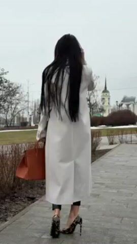 A woman standing on a paved walkway is featured in this video. The woman has long, dark hair and is wearing a light-colored, long coat, dark pants, and leopard-print high heels. She also has sunglasses on. She is holding a brown handbag in her left hand. She turns her head to the left, looking towards the camera, and then moves her hands in front of her. The background consists of a grassy area with bare trees, a church steeple in the distance, streetlights, and a building with a domed roof. The sky is overcast. The overall scene appears to be outdoors in an urban park-like setting on a cloudy day.