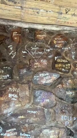 The video shows a close-up of a rock wall with graffiti. The graffiti is mostly written in white paint and includes names, dates, and other messages. There is a heart drawn around the names "Sonia" and "Victor." The text "PROMO 2023 DREAM" is visible on a rock in the middle of the frame. The wall also includes what appears to be bamboo trim above the stone and graffiti area. In the background someone mentions a spider.