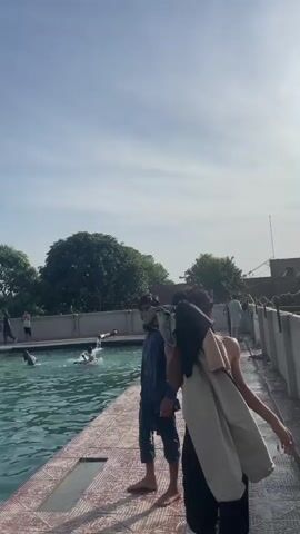The scene shows a group of people at a swimming pool on a sunny day. Some people are in the pool, swimming and splashing, while others stand or sit around the poolside. One man walks with clothes draped over his shoulder. Another man, dressed in a wet blue outfit, stands on the tiled edge of the pool. The pool is surrounded by a low wall, with trees visible in the background. The sky is mostly clear with some clouds.