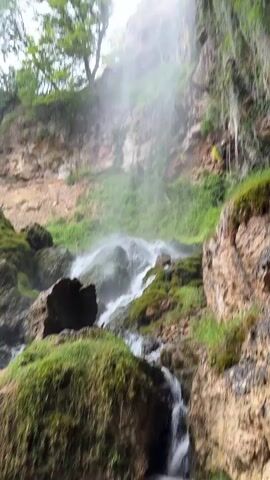 The video depicts a waterfall cascading down a rocky, moss-covered cliff face. Green trees and vegetation adorn the top of the cliff, while the sky above is overcast with grey clouds. The waterfall creates a mist as it plunges into the rocks below, and the overall scene evokes a sense of natural beauty and tranquility.