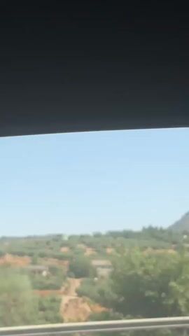 The video captures a view from a vehicle, looking out a window. The upper portion of the frame is dark, likely the interior of the vehicle. Below this, a bright blue sky is visible, with a distant hazy mountain range. The middle ground shows a landscape covered in green vegetation, possibly olive groves, with some scattered buildings. In the foreground, a blurred railing or barrier runs horizontally across the bottom of the frame. A grey signpost is visible in the middle distance. The scene appears to be a sunny day, likely in a rural or mountainous area.