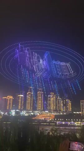 A drone light show is taking place at night over a cityscape. Hundreds of drones are illuminated with blue, purple, and white lights, forming intricate patterns in the sky. The drones create shapes of buildings, circular rings, and streams of what appears to be binary code. Below the drone display, a river reflects the city lights. The city skyline is composed of numerous tall buildings, many of which are illuminated with warm yellow and orange lights. In the foreground, a silhouette of trees is visible, and two hands are holding up smartphones, presumably to record the spectacle. The overall atmosphere is one of awe and technological advancement.