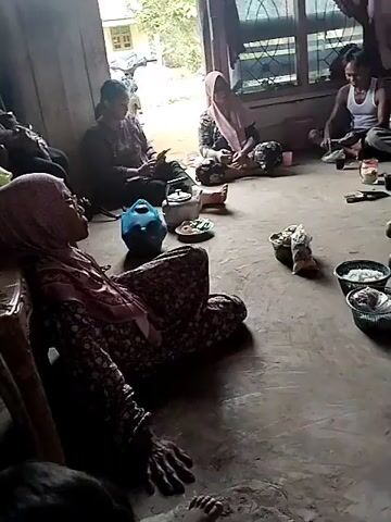 The video shows a group of people gathered in what appears to be a living room or communal area. Several individuals, predominantly women wearing headscarves, are seated on the floor, some leaning against furniture. One woman in the foreground, draped over a chair, is wearing a floral pattern headscarf and dress. In the background, other people are seated, and there are various objects on the floor including bowls of food, a blue water jug, and a kettle. The environment seems to be a modest dwelling with wooden walls and a large doorway or window leading to an outdoor area. The lighting suggests it is daytime, possibly with overcast conditions. The overall atmosphere appears to be one of a gathering, possibly a family or community event.