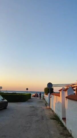 The video shows a sunset scene on a street leading to a beach. The sky is clear and blue with a hint of orange at the horizon. On the right side of the street is a white wall with a tiled roof, casting a shadow onto the street. On the left side, there is a trimmed hedge and a parked car. In the distance, at the end of the street, is the ocean and the beach. A group of people are walking towards the beach, some of them are near a horse-drawn cart. The overall atmosphere is peaceful and serene.