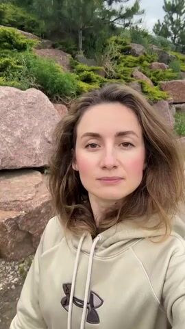 A woman wearing an Under Armour hoodie stands in front of a landscape with large rocks and greenery. She looks at the camera and turns her head to the side as a second woman, wearing a white t-shirt with a design of two wine glasses, enters the frame. The second woman has dark hair pulled back and wears a backpack. The first woman smiles into the camera.